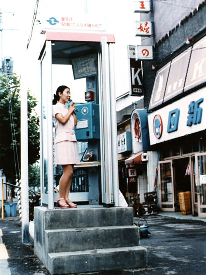 電話ボックスの大形青公衆電話で電話をする女性の写真